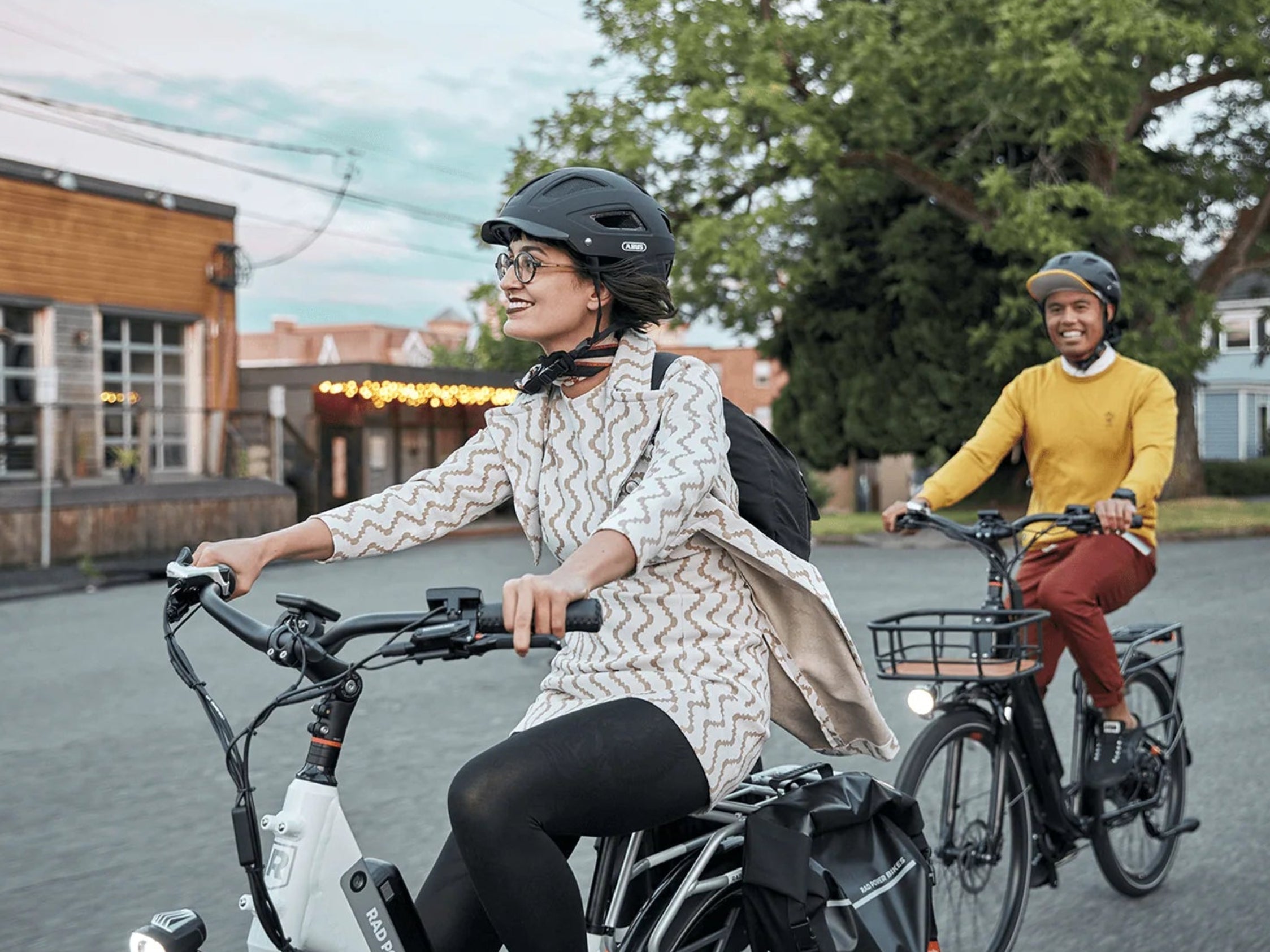 Two people riding electric bikes on a street with trees and buildings in the background.